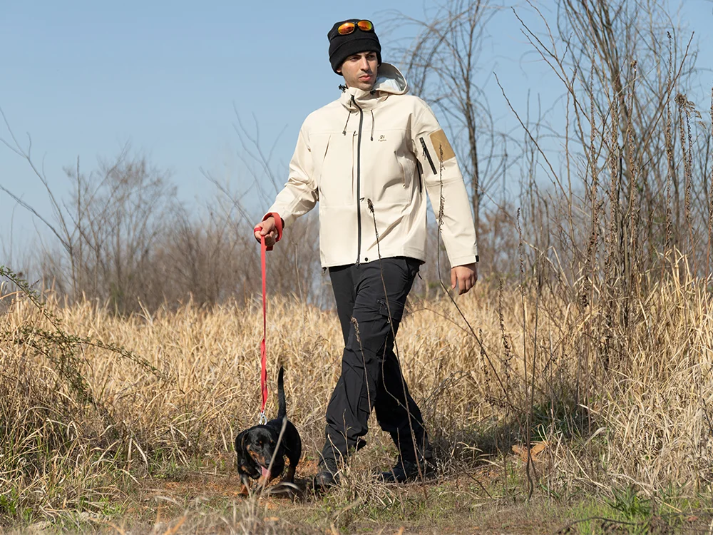 Man wearing a beige 3-in-1 waterproof outdoor jacket while walking a dog in a field, representing WHFrontend’s OEM outdoor clothing for all-weather performance.