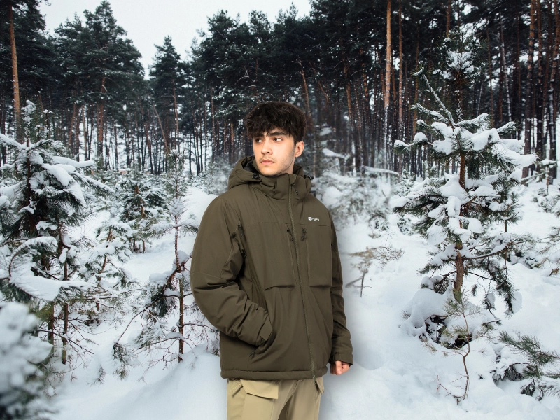 Man wearing an olive green insulated winter jacket in a snowy forest, representing WHFrontend’s warm and durable outerwear for cold climates.